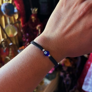 May include: A black woven cord bracelet featuring a central blue glass evil eye bead. The bead is round with a dark blue center, a white ring, and a light blue outer circle. Small silver-toned metallic beads flank the evil eye on both sides.