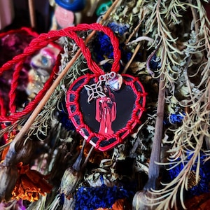 May include: A heart-shaped pendant featuring a black center with a red figure of a person in robes, framed by a red braided border. The pendant hangs from a red cord and includes two small silver-toned charms, one heart-shaped and one knot-shaped.