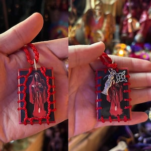 May include: A rectangular pendant featuring a central image of a hooded figure in a red robe holding a scythe, encased in a red cord lacing. The pendant is suspended from a red cord and includes a small silver-toned charm of a skeleton figure.