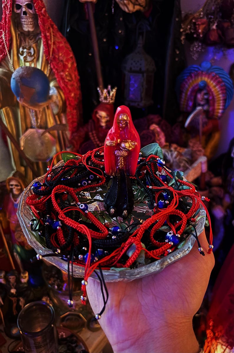 May include: A clear glass bowl filled with multiple braided red and black cord bracelets featuring small blue and white evil eye beads, topped with a small red and black figurine of a hooded figure with gold accents standing in the center.