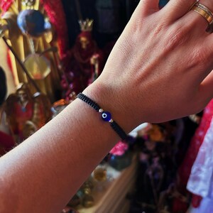 May include: A black woven cord bracelet featuring a central blue glass evil eye bead flanked by two small silver-toned metallic beads. The bracelet is constructed with a macrame-style knot pattern and is worn around the wrist.