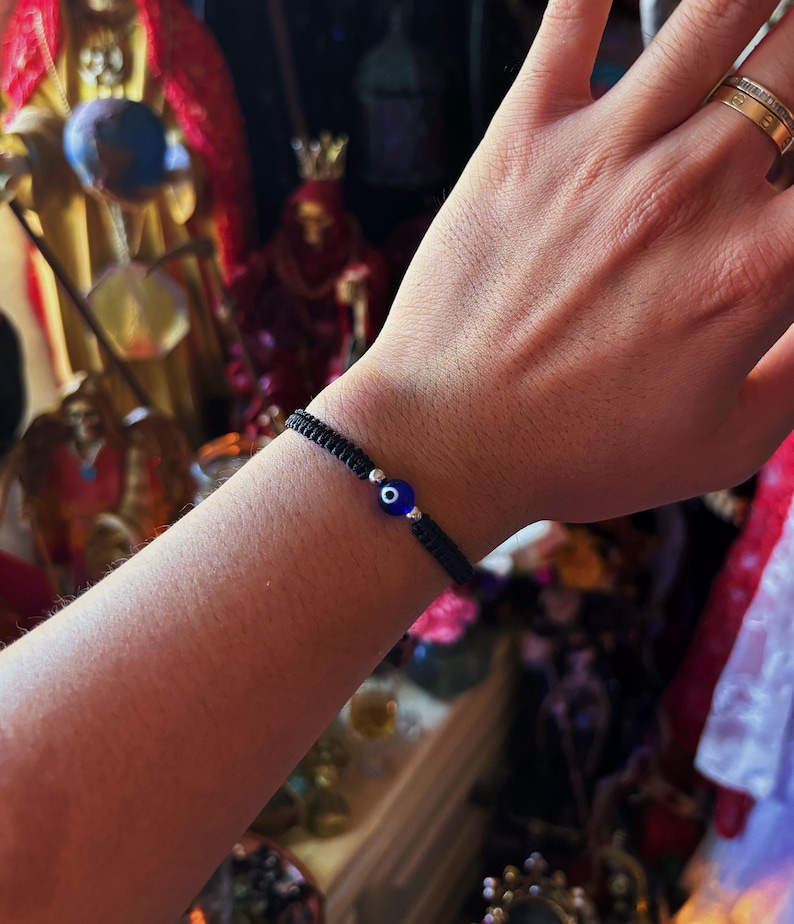 May include: A black woven cord bracelet featuring a central blue glass evil eye bead flanked by two small silver-toned metallic beads. The bracelet is constructed with a macrame-style knot pattern and is worn around the wrist.