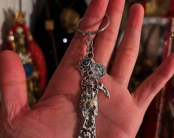 May include: A silver-toned keychain featuring a figure of Santa Muerte holding a scythe, accented with red stones and a white pearl. The keychain includes a small circular charm with a blue eye symbol and a round silver medal with religious text.