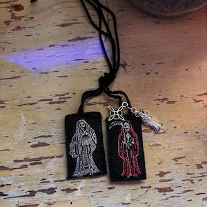 May include: Two rectangular black fabric patches attached to a black cord. One patch features an embroidered silver skeleton figure, while the other displays a red embroidered skeleton holding a scythe. A silver metal charm of a figure is attached.