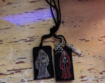 May include: Two rectangular black fabric patches attached to a black cord. One patch features an embroidered silver skeleton figure, while the other displays a red embroidered skeleton holding a scythe. A silver metal charm of a figure is attached.