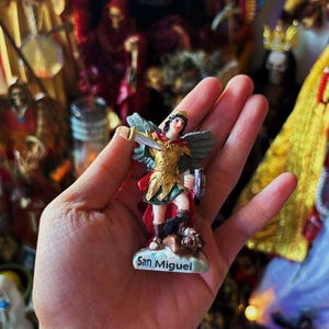 May include: A small figurine of San Miguel featuring a figure with wings, wearing gold armour and a red cape, holding a sword and a shield while standing over a dark figure. The base of the figurine is white and clearly displays the text San Miguel.