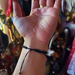 May include: A black cord bracelet featuring an adjustable sliding knot closure mechanism. The bracelet is constructed from thin, durable black string and includes two small metallic beads at the ends of the adjustable pull cords for a secure fit.