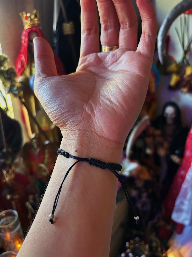 May include: A black cord bracelet featuring an adjustable sliding knot closure mechanism. The bracelet is constructed from thin, durable black string and includes two small metallic beads at the ends of the adjustable pull cords for a secure fit.