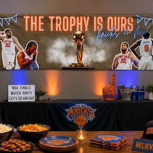 Op de afbeelding: Een New York Knicks themafeest opstelling met een grote banner met de tekst THE TROPHY IS OURS en knicks in five, een basketbaltrofee, spelersafbeeldingen, een lichtbak met feesttekst, snacks, borden en een zwarte pet met het teamlogo.