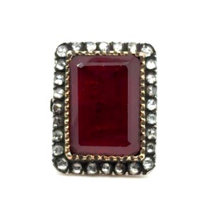 May include: A rectangular ruby-colored gemstone ring with a gold-colored border. The ring is surrounded by small, clear gemstones set in a silver-colored metal. The ring is a statement piece.