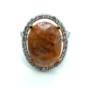 Può includere: Anello in argento con una pietra preziosa ovale marrone chiaro. La pietra è circondata da un'aureola di piccole pietre trasparenti. Il design dell'anello è classico ed elegante, adatto a varie occasioni.