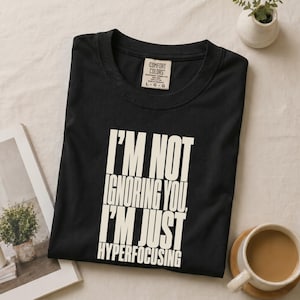 Op de afbeelding: Een zwart T-shirt met korte mouwen, een ronde hals en een Comfort Colors merklabel. Op de voorkant staat de witte tekst: I'M NOT IGNORING YOU, I'M JUST HYPERFOCUSING. Het T-shirt is gemaakt van katoen en heeft maat large, zoals aangegeven op het label.