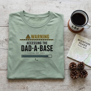 May include: A sage green crew neck t-shirt featuring a black and gold graphic that reads WARNING ACCESSING THE DAD-A-BASE loading dad jokes with a progress bar and a blinking cursor icon. The design includes a yellow warning triangle symbol.