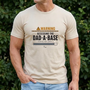 May include: A beige short-sleeved t-shirt featuring a graphic with a yellow warning triangle and the text: WARNING ACCESSING THE DAD-A-BASE loading dad jokes... along with a horizontal loading bar and a blinking cursor underscore symbol.
