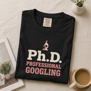 May include: A black crew neck t-shirt featuring a light pink microscope icon above the white text Ph.D. and the pink text PROFESSIONAL GOOGLING. The shirt has a Comfort Colors brand label on the inside collar indicating a size large.