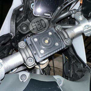 Op de afbeelding: Close-up van een motorstuur met een zwarte telefoonhouder en een tablet. De houder is met bouten bevestigd. De motorfiets heeft een grijs en wit kleurenschema. De tekst "H3D" is zichtbaar.