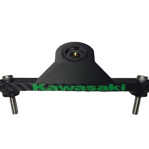 Op de afbeelding: Een zwart motorfietsonderdeel met het groene woord "Kawasaki" erop gedrukt. Het onderdeel heeft een koolstofvezelpatroon en twee zilveren bouten. Het heeft een bevestigingspunt aan de bovenkant en twee bouten aan de onderkant.