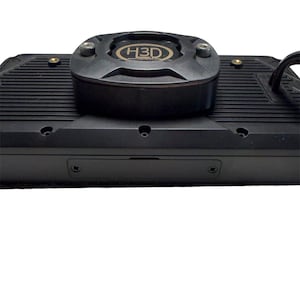 May include: A black rectangular electronic cooling device featuring a central circular fan housing with the text H3D printed in gold. The unit has a ribbed surface texture, multiple visible screw points, and a black power cable extending from the side.