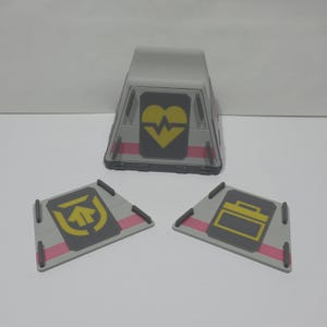 May include: A grey and clear plastic structure featuring a yellow heart symbol and pink accents. Three grey, triangular pieces with yellow symbols and pink accents are also present. The objects appear to be part of a toy or model.