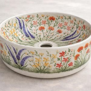 May include: A round, white ceramic sink basin with a floral design. The basin features a hand-painted pattern of colorful flowers, including red, yellow, and purple blooms, set against a white background. The sink has a central drain hole.