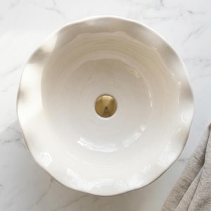 May include: A round, off-white ceramic vessel sink featuring a smooth, glossy finish and a distinctively wavy, scalloped rim. The basin includes a centred, circular drain cover with a brushed gold metallic finish, viewed from directly above.