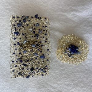 Könnte beinhalten: Ein goldfarbenes Drahtarmband und eine passende Brosche. Das Armband ist rechteckig und mit blauen Steinen und Perlen gefüllt. Die Brosche ist kreisförmig mit einer Ansammlung blauer Steine in der Mitte. Beide Teile liegen auf einer weißen Oberfläche.