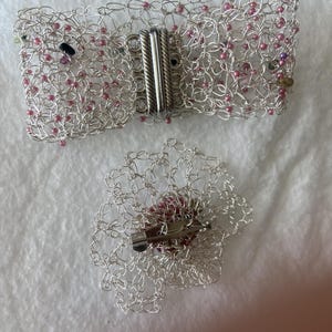 Könnte beinhalten: Ein silberfarbenes Drahtgeflechtarmband mit Magnetverschluss und eine passende blütenförmige Brosche. Das Armband ist mit kleinen rosa Perlen verziert. Die Brosche hat eine Nadelrückseite und einen zentralen rosa Perlencluster.
