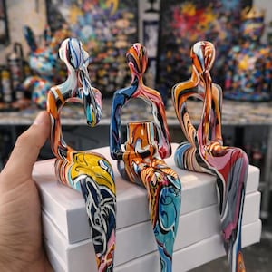 Puede incluir: Tres esculturas abstractas de figuras sentadas con patrones coloridos y arremolinados. Las esculturas se sostienen sobre una pila de cajas blancas. Las figuras tienen un diseño artístico moderno con una variedad de colores, incluyendo naranja, azul, amarillo y rosa.