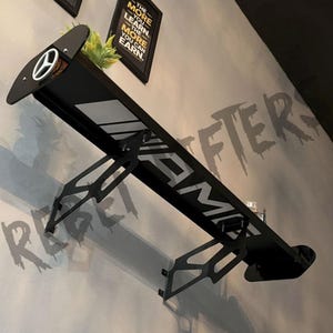 May include: Black metal wall shelf designed to resemble a car spoiler, featuring the AMG logo in white. The shelf has a white stripe and a Mercedes-Benz logo. The wall behind the shelf has the words "REFTERS" and "THE MORE YOU LEARN, THE MORE YOU CAN EARN."