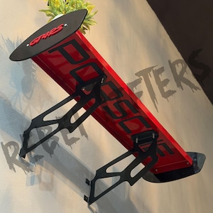 May include: A red and black car spoiler with the word "POSSE" in large black letters. The spoiler has a black top piece with the word "GRES" in red. The spoiler is mounted on a wall with black brackets.