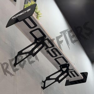 May include: A wall-mounted shelf designed to resemble a black and white automotive rear spoiler. The shelf features the word PORSCHE in bold black lettering on the white surface and on the black side end plates, supported by two black metal brackets.