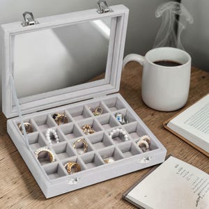 May include: A white ceramic mug with a black handle and the text "But First, Coffee" in a bold, black font. The mug is set against a blurred background, suggesting a cozy setting.