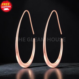 May include: A pair of rose gold-toned hoop earrings featuring an elongated, teardrop-shaped silhouette with a smooth, polished finish. The earrings have a continuous, open-loop design that tapers to a point at the top, creating a sleek, curved form.