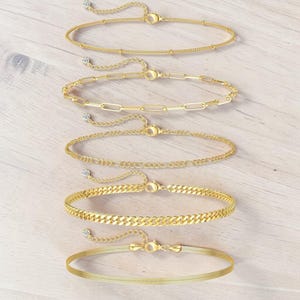 May include: Several gold-coloured anklets in different styles, such as beaded, paperclip, curb, and herringbone. Each anklet has an adjustable chain and a small, sparkling detail. The anklets are presented on a light wood-grain surface.