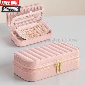 May include: A rectangular light pink jewelry box with a quilted top and gold-tone double zipper closure. The open interior features a clear window lid, multiple ring rolls, and small compartments for organizing earrings and rings.
