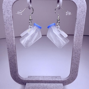 Könnte beinhalten: Ein Paar transparente, geometrische Ohrringe mit blauen Akzenten. Die Ohrringe sind an silberfarbenen Fischhaken-Ohrdrähten befestigt. Die Ohrringe werden auf einem gesprenkelten, hellgrauen Ohrringhalter präsentiert.