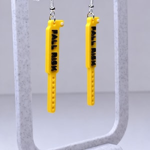 May include: A pair of yellow earrings with the text "FALL RISK" in black. The earrings are rectangular with a series of small holes along the bottom edge. They hang from silver-coloured hooks and are displayed on a grey stand.