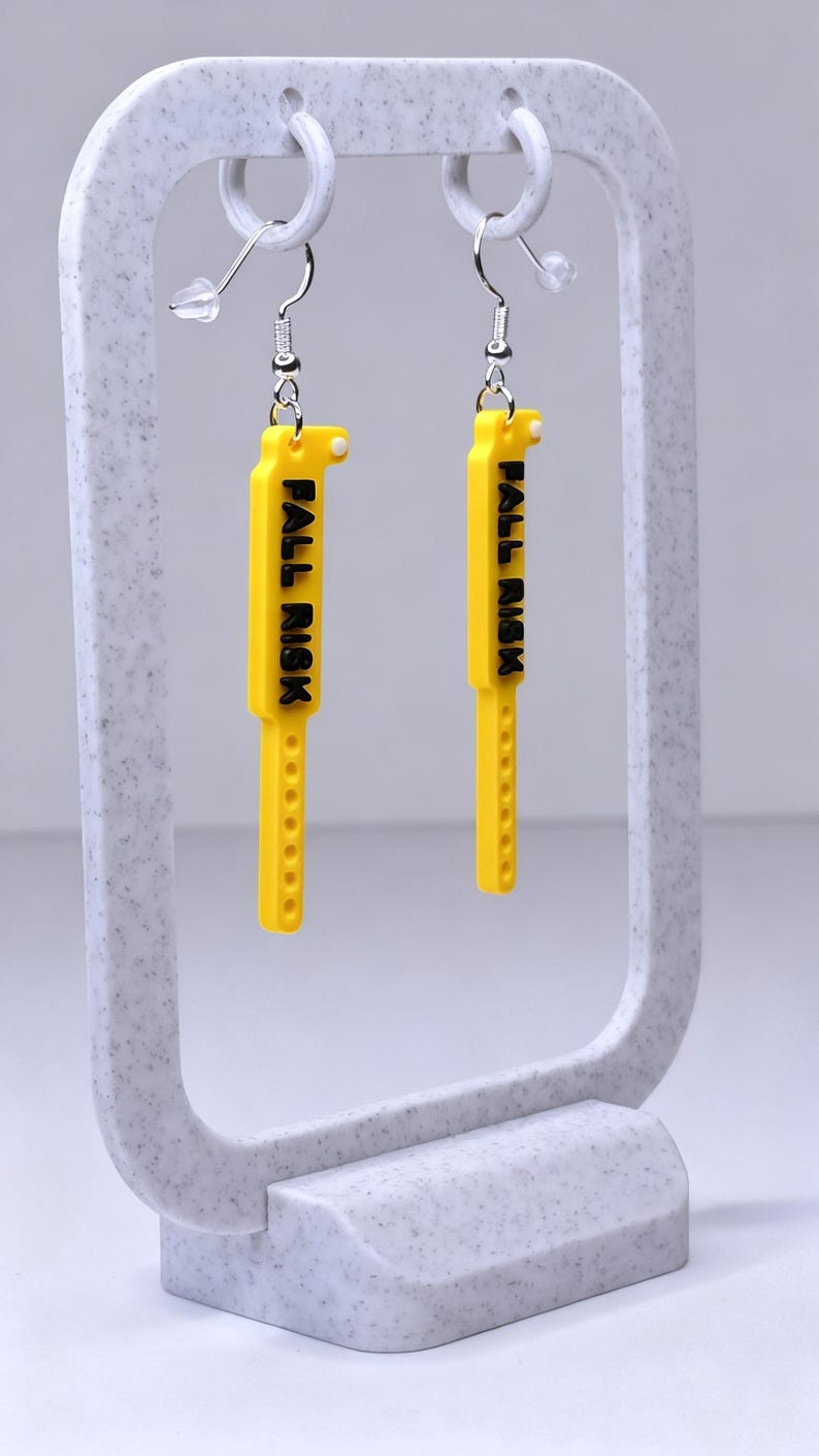 May include: A pair of yellow earrings with the text "FALL RISK" in black. The earrings are rectangular with a series of small holes along the bottom edge. They hang from silver-coloured hooks and are displayed on a grey stand.