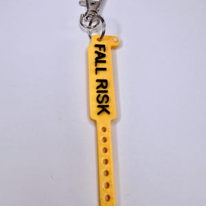 May include: A yellow, rectangular keychain with the words "FALL RISK" printed in black. The keychain has a series of small holes and a metal clasp for attachment. The design is simple and functional, suitable for medical or care settings.