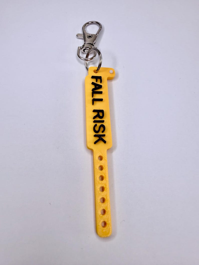 May include: A yellow, rectangular keychain with the words "FALL RISK" printed in black. The keychain has a series of small holes and a metal clasp for attachment. The design is simple and functional, suitable for medical or care settings.
