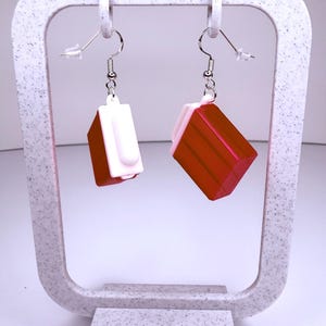 Puede incluir: Un par de pendientes geométricos rojos y blancos. Cada pendiente presenta una base roja angular con una parte superior rectangular blanca. Los pendientes cuelgan de ganchos plateados. Los pendientes se exhiben en un soporte blanco moteado.
