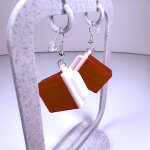May include: Earrings with a geometric design. The earrings are brown and white, with a rectangular shape. They are hanging from a white speckled earring display stand. The earrings have silver-colored hooks and small clear beads.