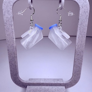 May include: A pair of clear, geometric earrings with blue accents. The earrings are suspended from silver-colored hooks and displayed on a speckled, light purple earring stand. The earrings have a unique, modern design.