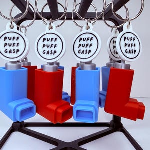 May include: Keychains featuring miniature inhaler designs in red and blue, each with a white tag printed with "PUFF PUFF GASP" in a circular design. The keychains are attached to silver rings and displayed on a black stand.