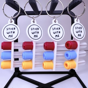 Puede incluir: Cuatro llaveros con el texto "STICK WITH ME" en etiquetas circulares blancas. Cada llavero tiene un anillo de metal y está unido a un tubo transparente con tapas rojas, azules y amarillas. Los llaveros se exhiben en un soporte negro.