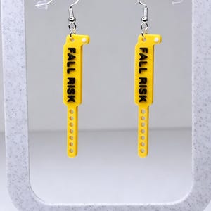 May include: A pair of yellow earrings with the text "FALL RISK" in black. The earrings are rectangular with a rounded top and a series of small holes along the bottom. They hang from silver-colored ear wires.