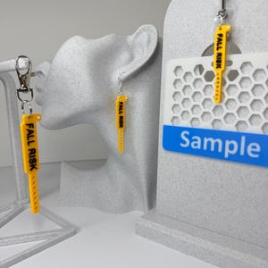May include: Yellow plastic accessories shaped like hospital wristbands with the text FALL RISK printed in black. The set includes a keychain with a metal clasp, a pair of earrings with silver-toned hooks, and a badge reel attachment for an ID holder.