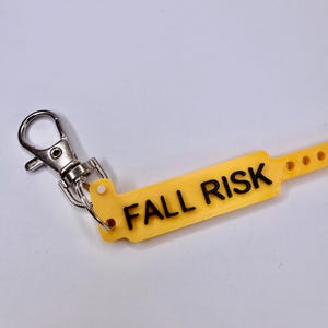 May include: A yellow tag with the words "FALL RISK" in black lettering, attached to a silver metal clip. The tag has a series of small holes along one edge. The background is a plain white surface.