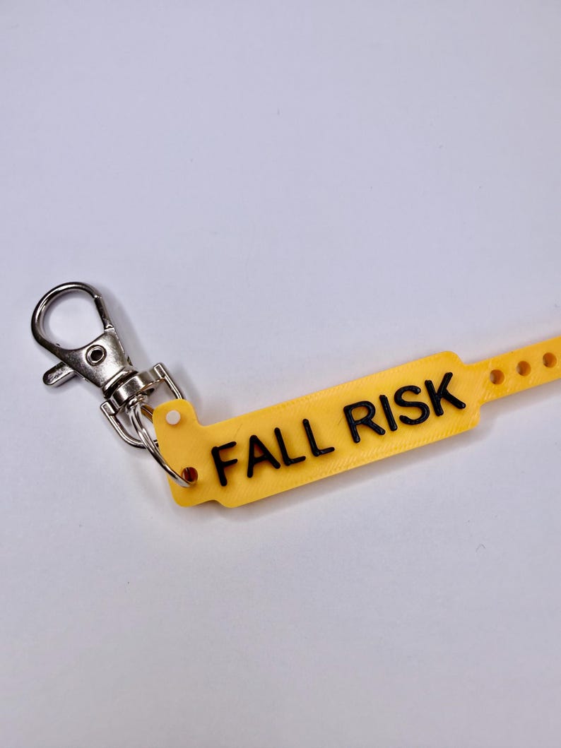 May include: A yellow tag with the words "FALL RISK" in black lettering, attached to a silver metal clip. The tag has a series of small holes along one edge. The background is a plain white surface.