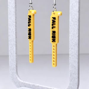 May include: Yellow earrings with the text "FALL RISK" in black lettering. The earrings are rectangular with a series of small holes along the bottom edge. They hang from silver-coloured ear wires and are displayed on a white stand.
